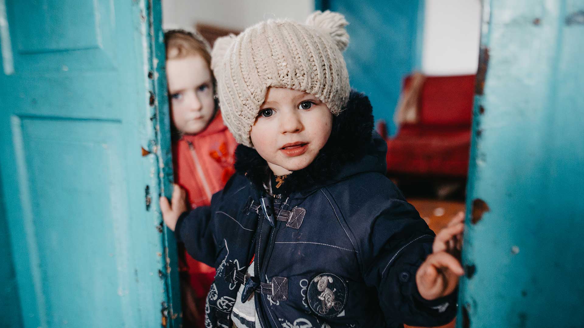 Young child in winter clothing and a knitted hat standing in a doorway, with another child leaning against the door in the background.